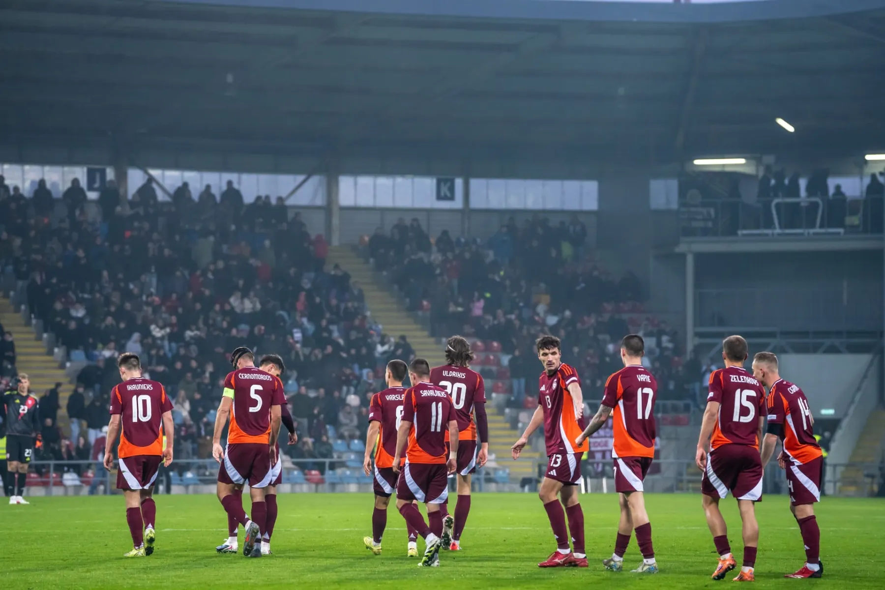 Latvijas izlases futbolisti bordo formās iet pa laukumu stadionā pilnu tribīņu priekšā